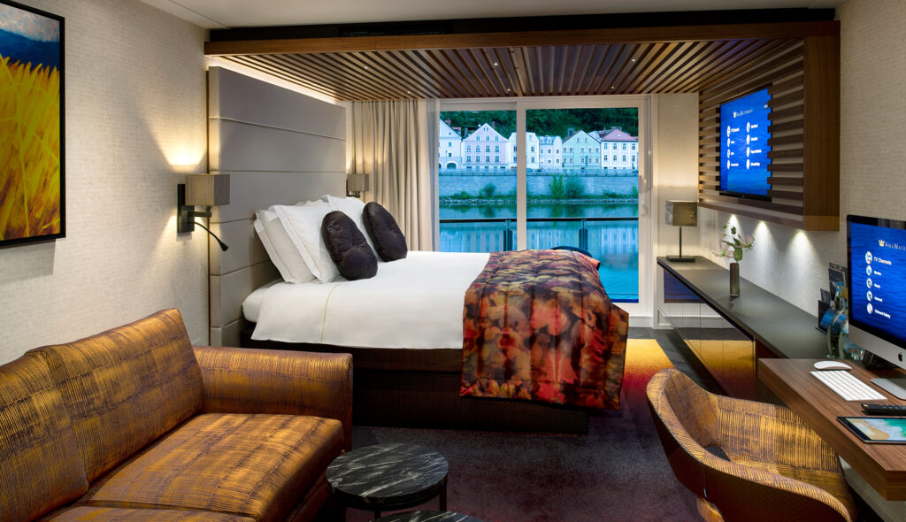 Suite aboard the AmaMagna with a large bed, a sofa, and a window overlooking the riverbank